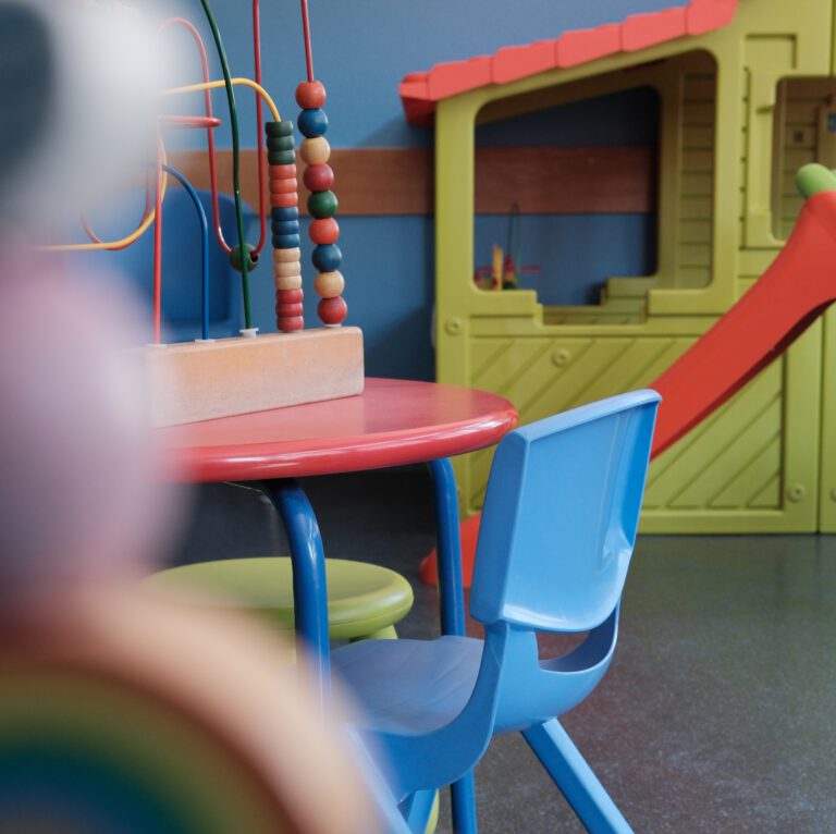 Das Wartezimmer unserer Kinderarztpraxis mit einem blauen Stuhl vor einem Tisch und einem Spielhaus im Hintergrund
